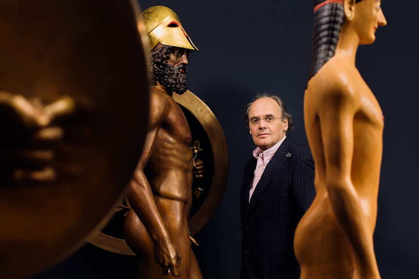 A white man in his 60s wearing glasses and suit stands between two golden statues