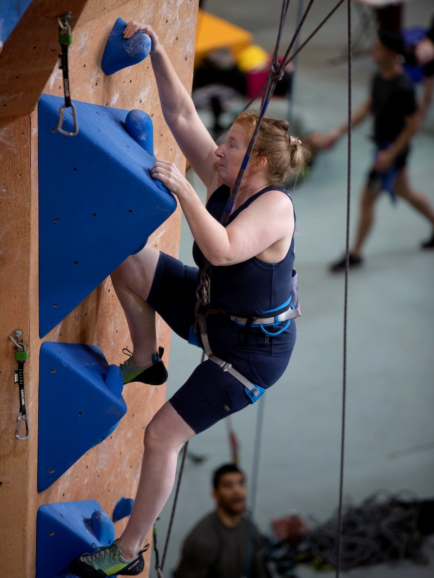 Julia Postma reaches up with her hands while climbing blue blocks on an interior wall.