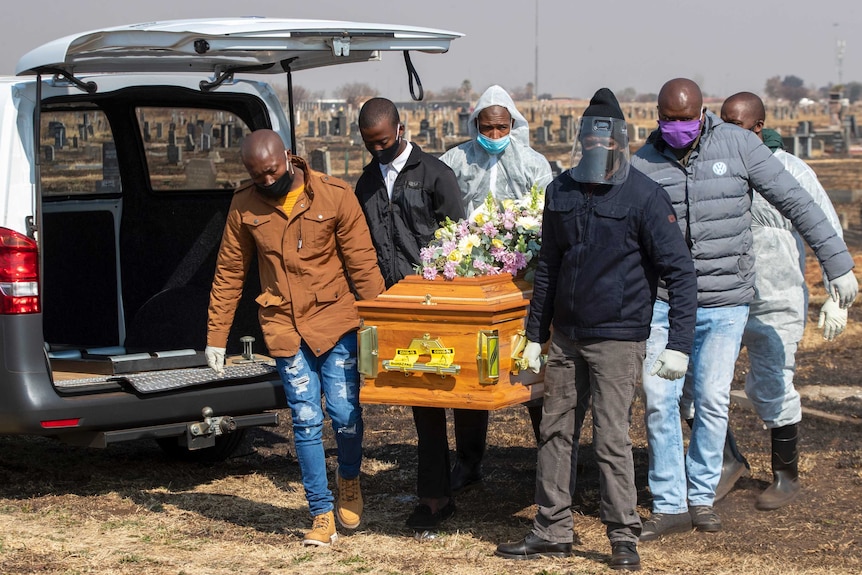 Six men wearing face masks and protective gear carry a coffin from the rear of a van.