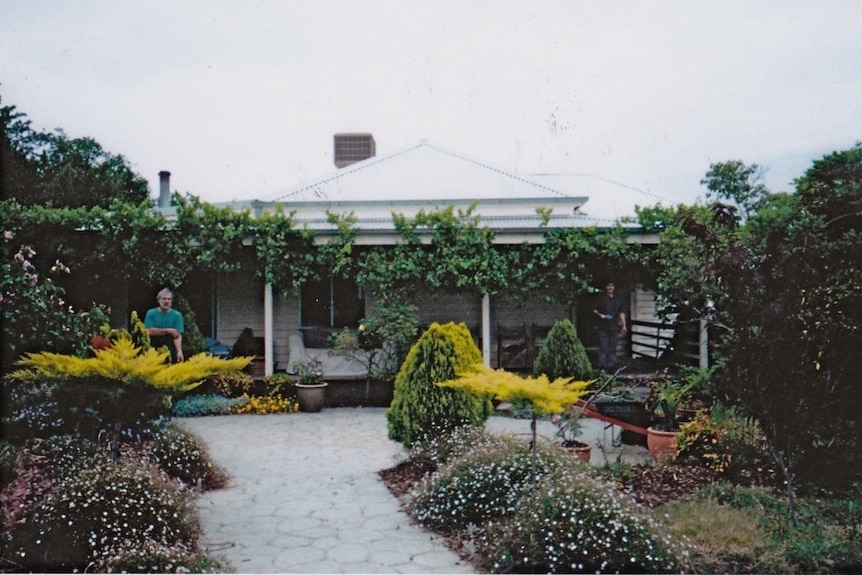 An old farmhouse, surrounded by garden, with two people standing outside it.