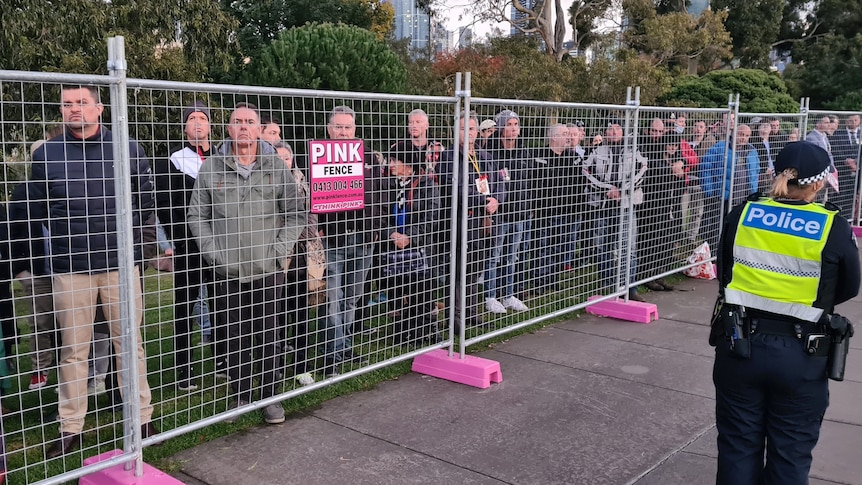 A crowd of people watches a dawn service from outside a temporary fence.