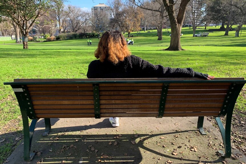 A woman photographed from behind sits on a bench in a park.