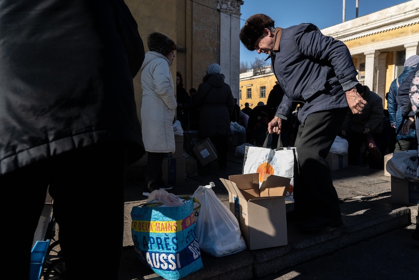 An old man leans over a shopping bag, one of several sitting on a footpath outside a building