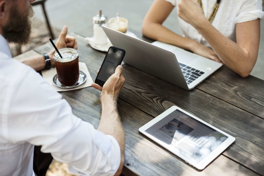 Two people sit at a cafe table and use their phone, laptop and iPad.