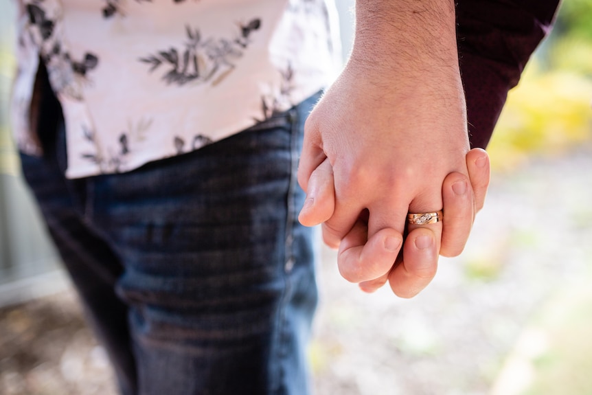 Close up of two men holding hands, one of them has a wedding ring.
