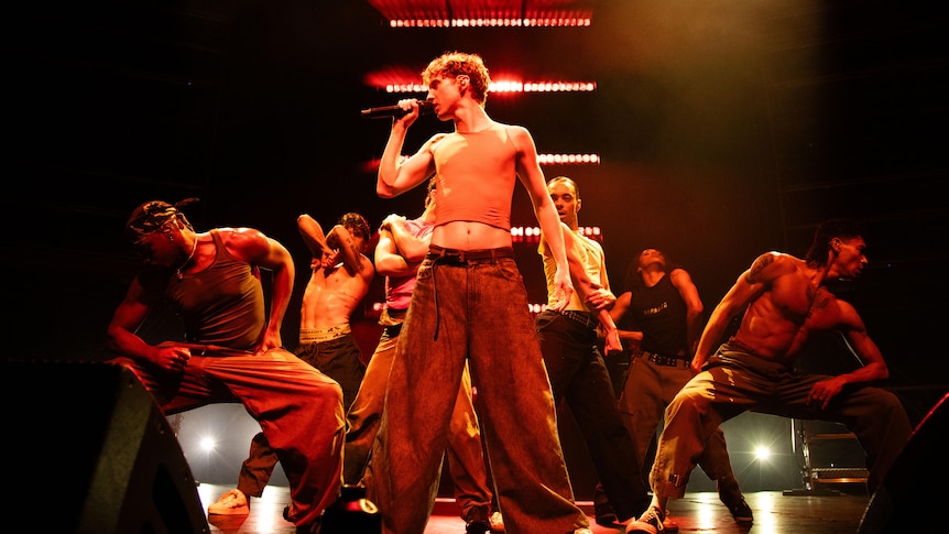Shirtless Troye Sivan singing on stage flanked by six male dancers.