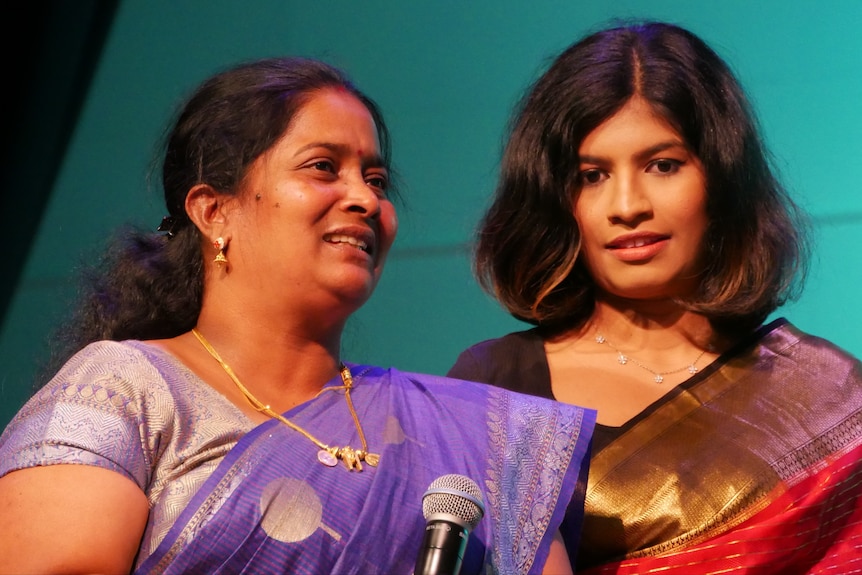 Priya holding a microphone wearing purple, Vashini wearing red.