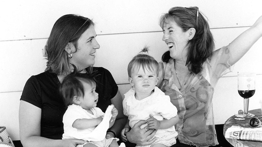 Black and white image shoes two young mothers nursing toddlers and having a drink at a Melbourne Cup Day party.