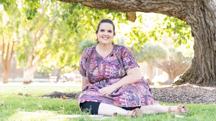 a smiling woman with dark hair and a purple Indigenous print dress sits on the grass beneath the limb of a tree