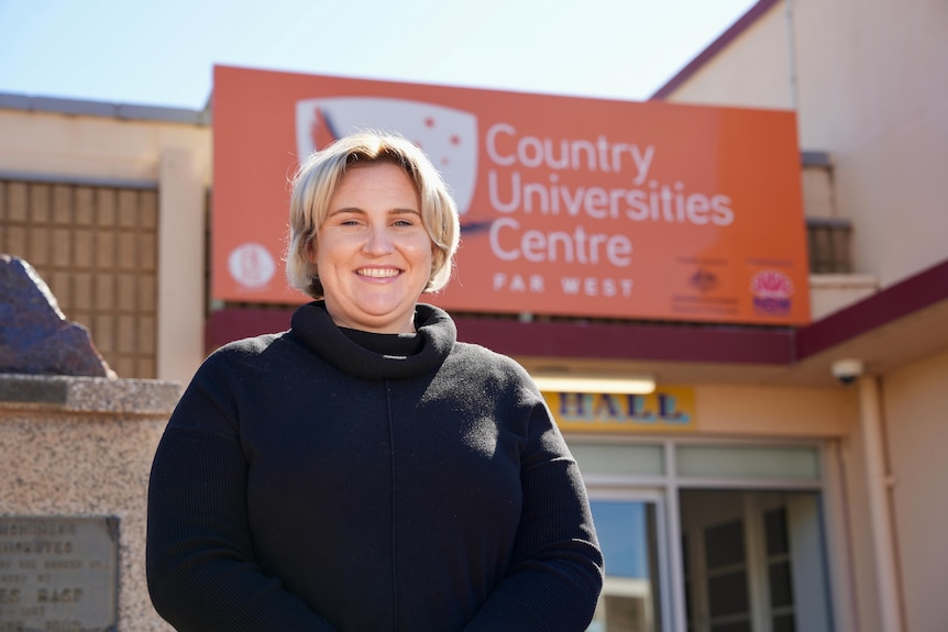 A blonde woman in a black turtleneck smiling outside a building with an orange sign.