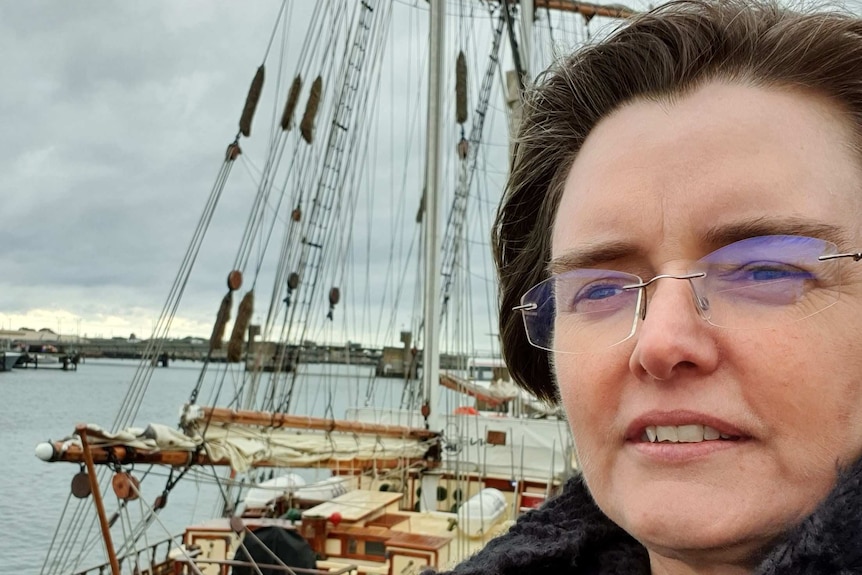 A woman with glasses looks out to sea with an old sail ship in the background.
