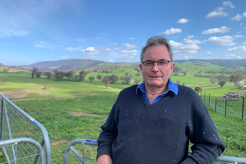 A man leaning on a gate with green paddocks in the background.