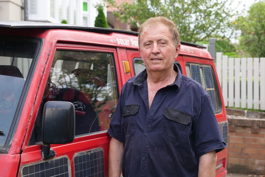 a man in mechanics clothes smiles standing next to a red van