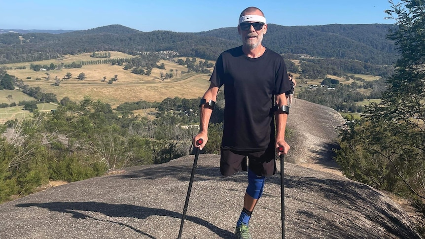 A man with one leg holding crutches stands on top of a mountain. He's wearing a dark t-shirt and shorts and sunglasses. 