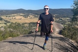 A man with one leg holding crutches stands on top of a mountain. He's wearing a dark t-shirt and shorts and sunglasses. 