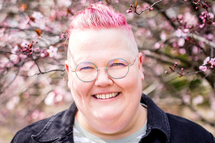 Jasper has short pink hair and thin green circular glasses. They stand against a pink cherry blossom tree background smiling.
