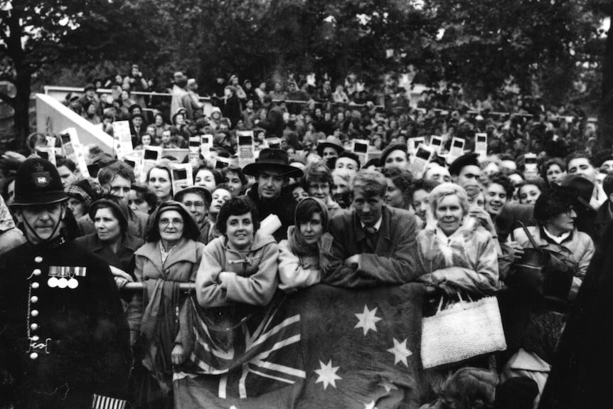 A black and white photo of a crowd, all smiling and looking happy. The people at the front are holding a large Australian flag.