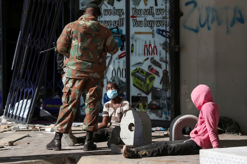 A soldier stands next to a person in a pink hoody who is sitting on the ground