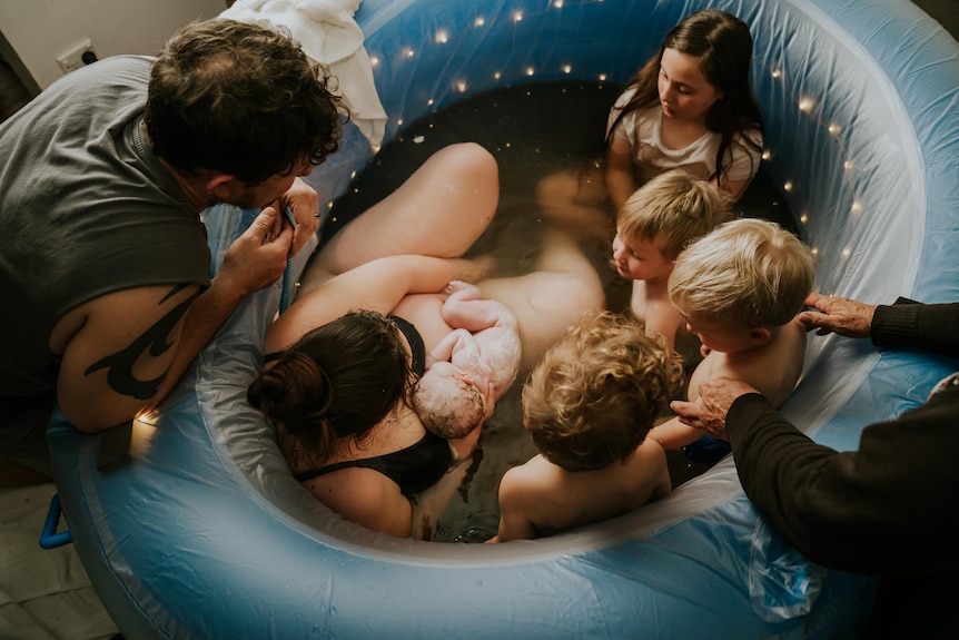 A woman lies in a birthing pool holding a newborn baby, surrounded by children, with a man leaning over them