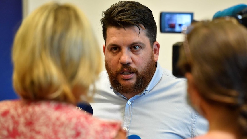 A bearded man speaks to two female journalists in a room.