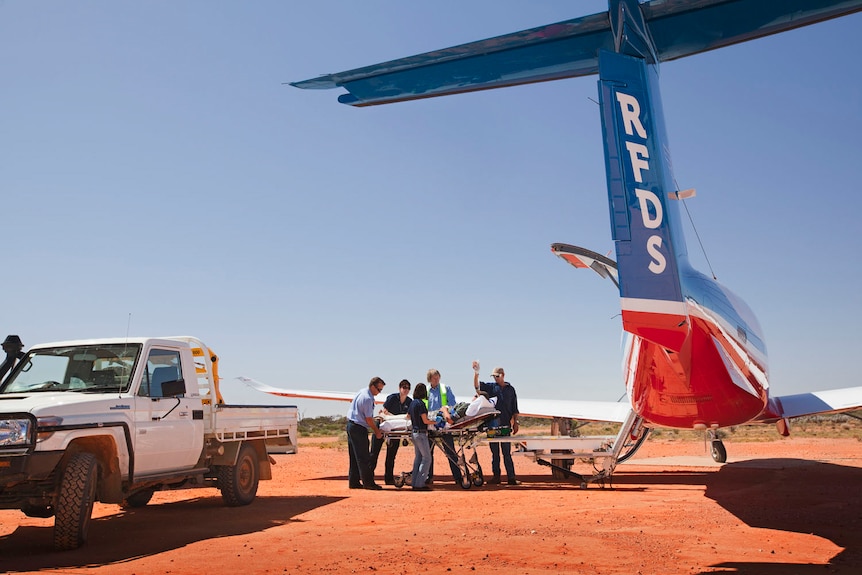 A patient is loaded into a plane parked on a red dirt runway.