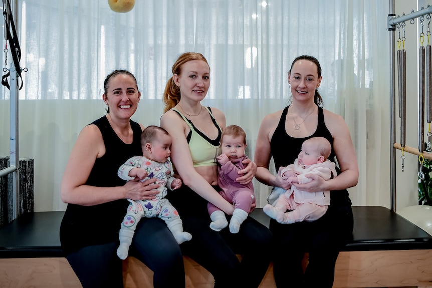 Three women sit on a reformer pilates bed holding their babies 