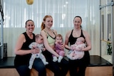 Three women sit on a reformer pilates bed holding their babies 