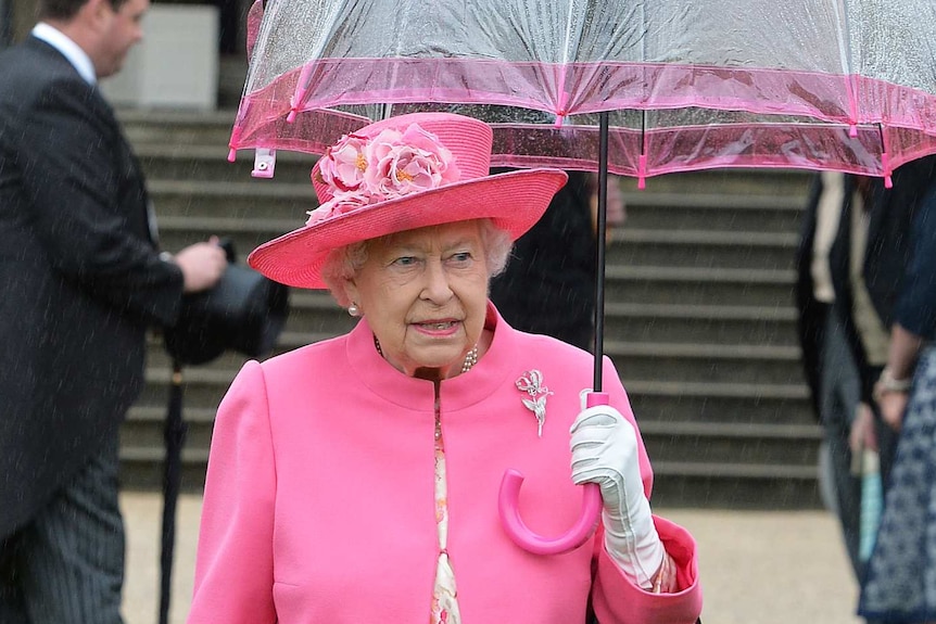 Queen Elizabeth II in pink walks under an umbrella in the garden of Buckingham Palace.