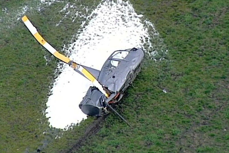 The crashed helicopter, missing its tail rotor, surrounded by fire retardant foam on the ground.