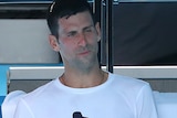 Novak Djokovic sits on a chair holding a bottle of juice, with his left leg folded over his right
