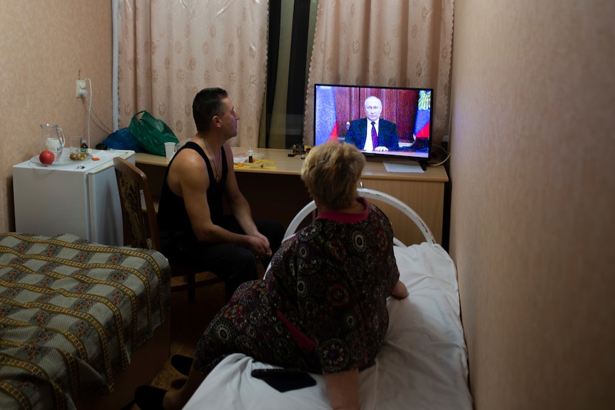 People in a small room watch a television showing Vladimir Putin. 