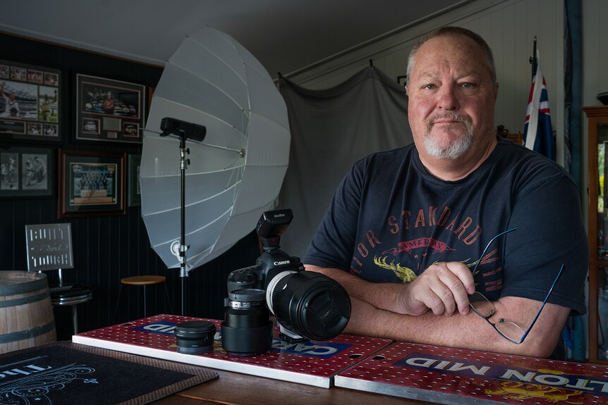 A man leans against a bar with a large camera and photography equipment in the background