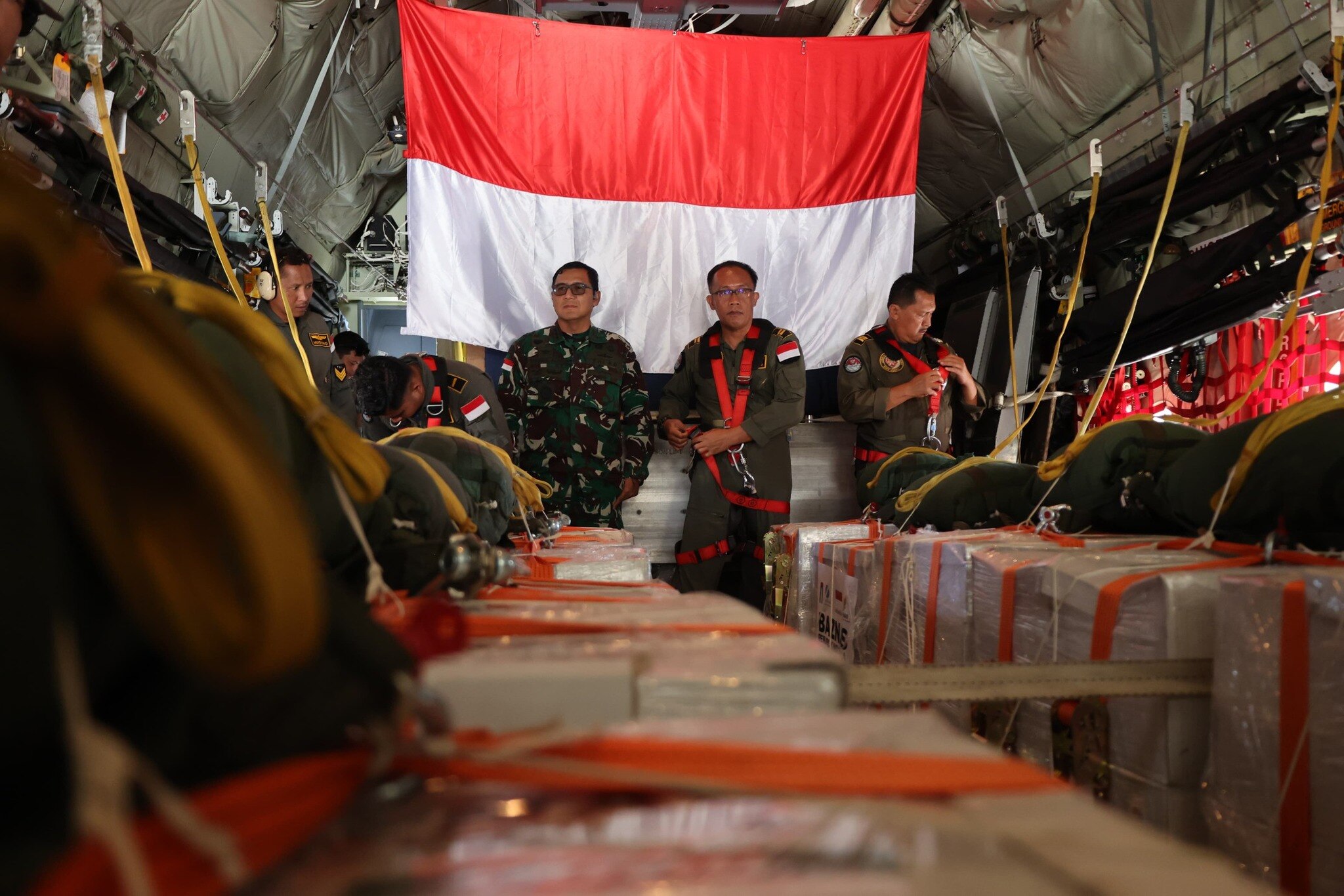 Three Indonesian army members standing behind boxes with Indonesian flag in the background.