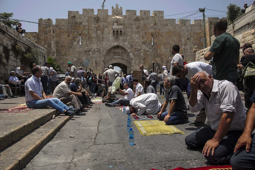 Men sit and kneel praying in front of a large stone wall.