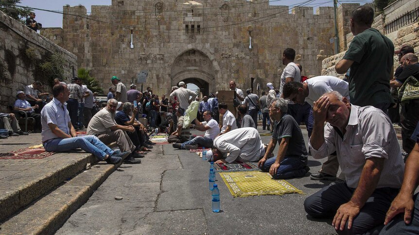 Men sit and kneel praying in front of a large stone wall.