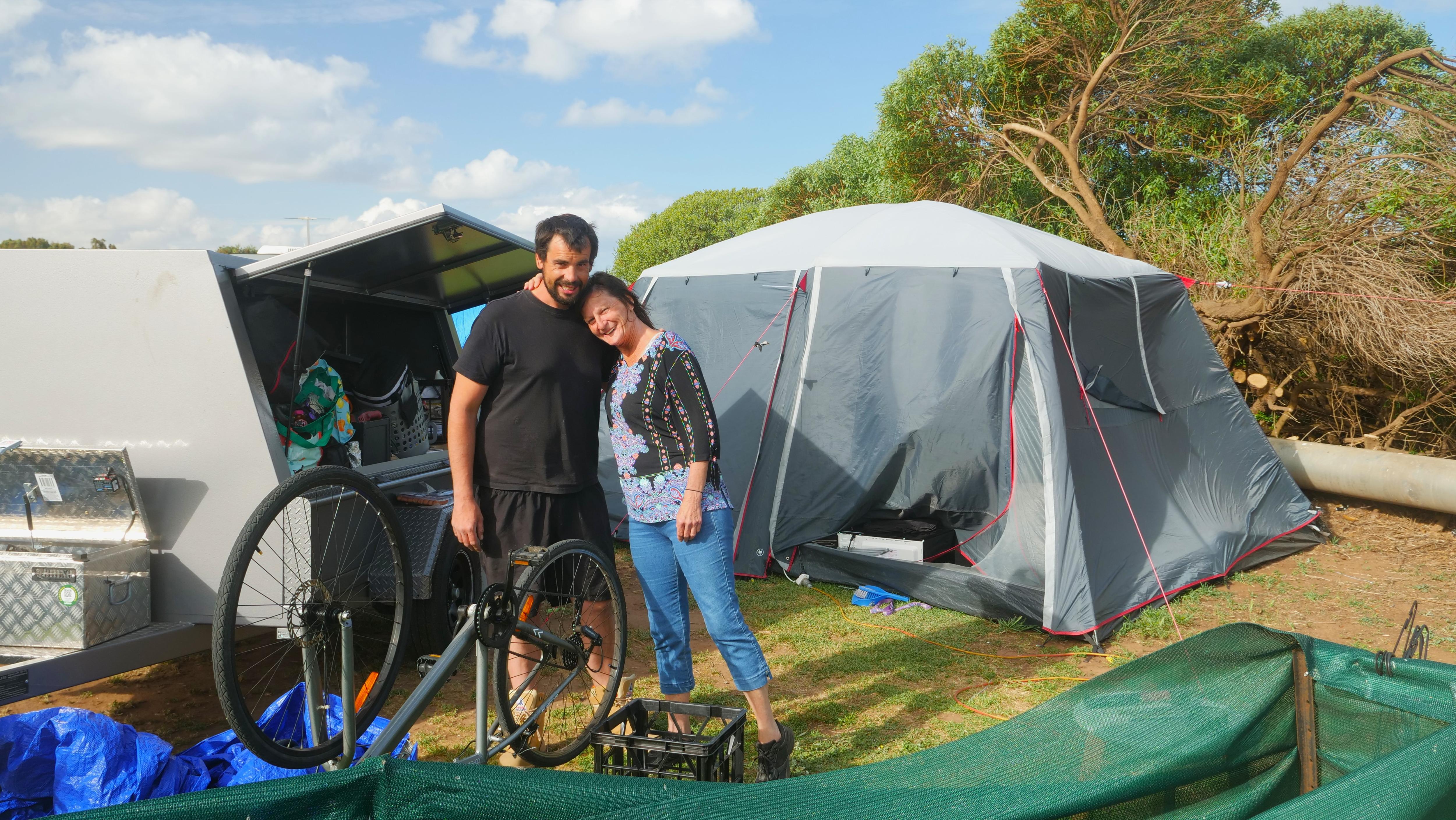 Man in black shirt hugging his mum, lady in jeans with colourful top, packing up gear in front of grey tent and trailer 