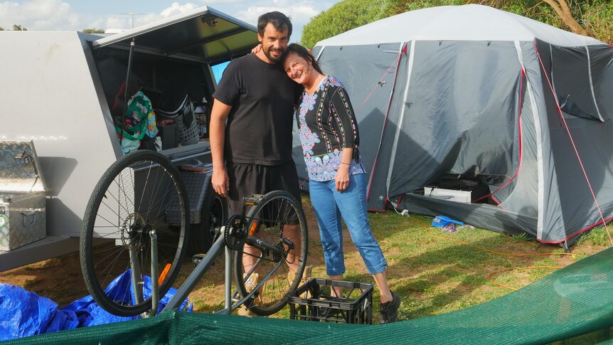 Man in black shirt hugging his mum, lady in jeans with colourful top, packing up gear in front of grey tent and trailer 