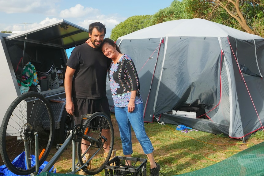 Man in black shirt hugging his mum, lady in jeans with colourful top, packing up gear in front of grey tent and trailer 