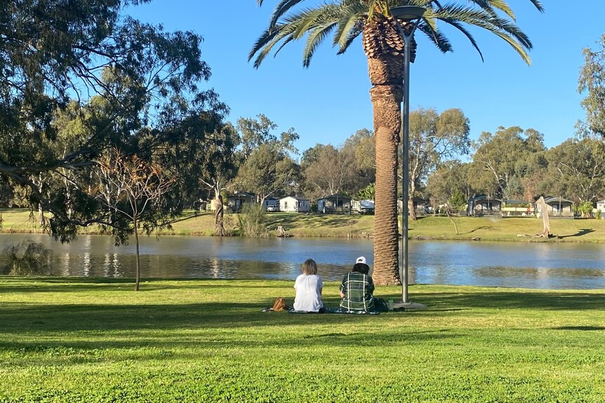 Two people sit side by side on grass by the banks of the Murray River