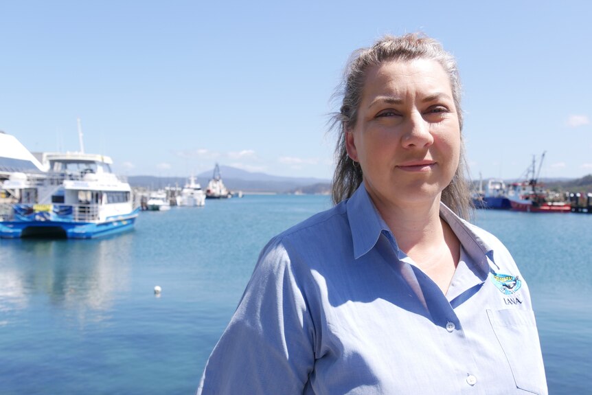 A woman with blonde hair in a blue shirt outside. Boats and water behind her.