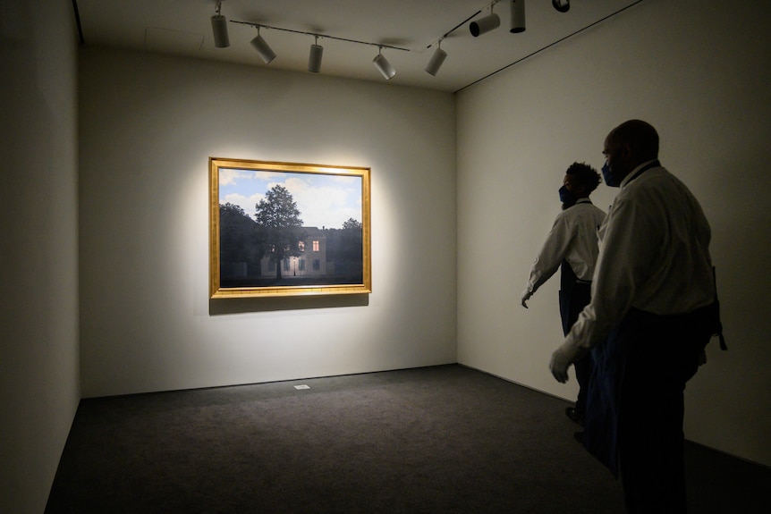 Two men walk into a dark room with a painting displayed under lights on the far wall.