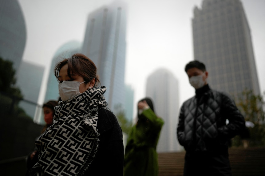 people wearing facemasks walk outdoors with buildings behind them