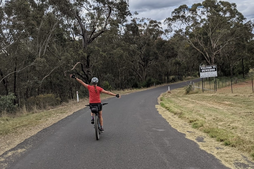 A woman on a bike with both her arms out on a road.