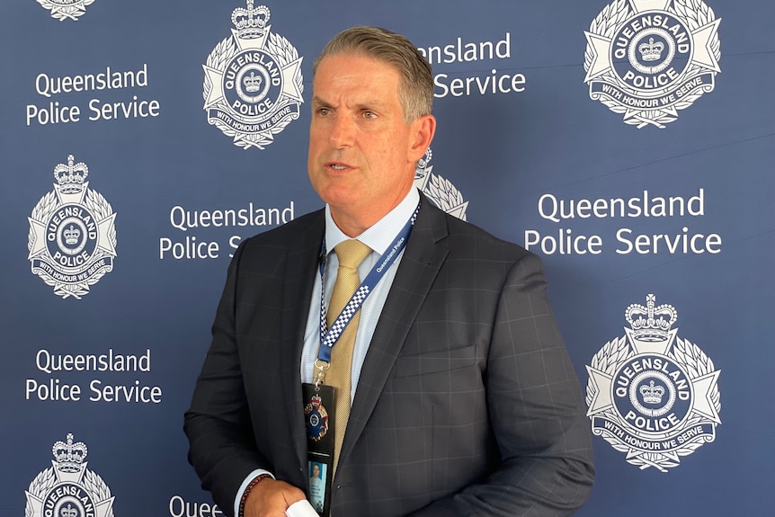 A man in a suit looks off camera, he is standing in front of a police backdrop