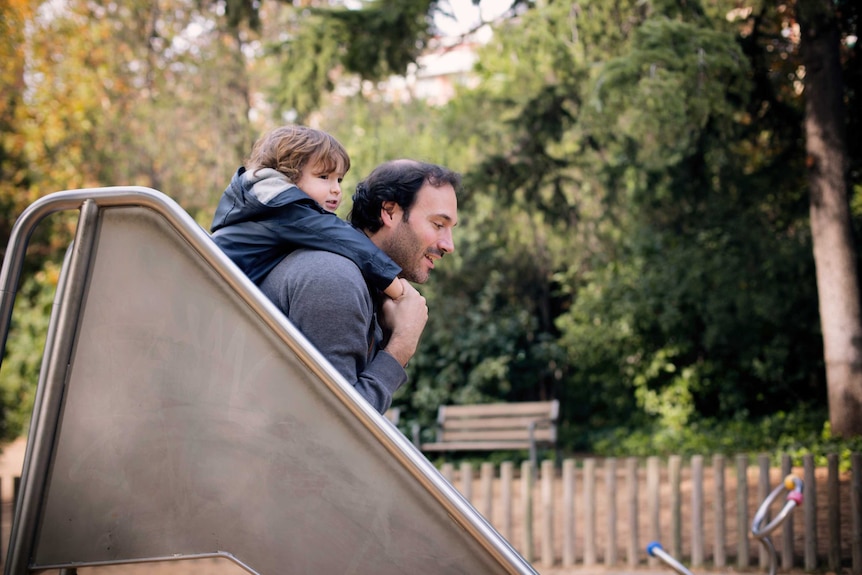 A father and son go down a slide together at a playground.