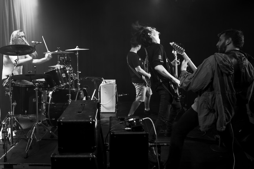 Monochrome of a four-piece rock band playing at  full noise on a stage.