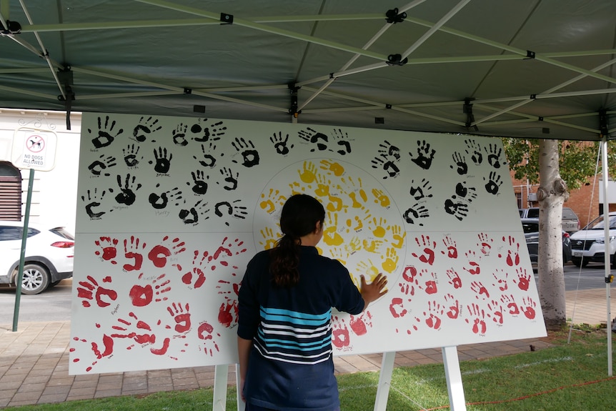 An Indigenous teenage girl wearing a black and blue striped jumper puts her hand print on an Aboriginal flag