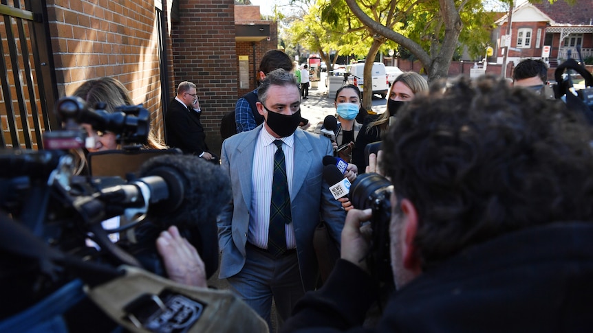 man walks through multiple journalists and camera's to doorway wearing a mask
