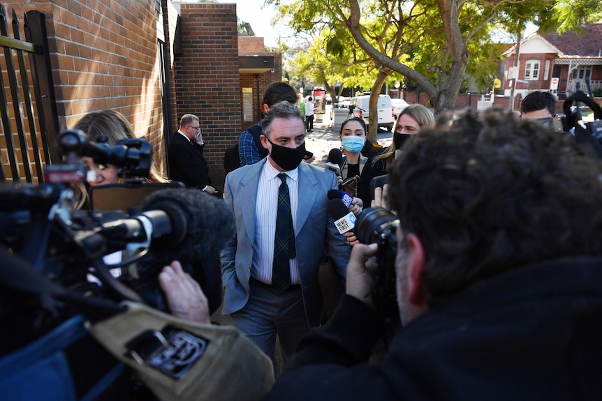 man walks through multiple journalists and camera's to doorway wearing a mask