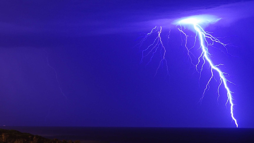 A massive bolt of lightning cuts through a blue sky, with part of the coast lit up by it.